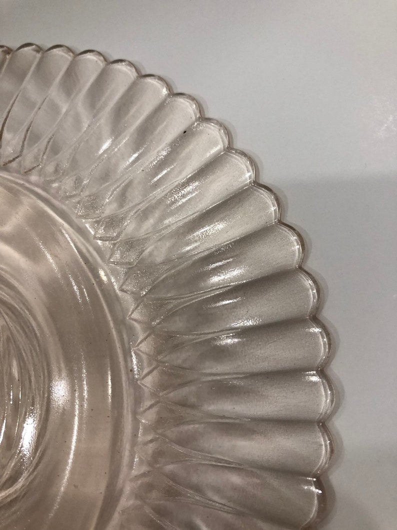 Puede incluir: Primer plano de un plato de vidrio transparente con borde festoneado. El plato presenta un dise&ntilde;o texturizado y acanalado que se extiende desde el centro hasta el borde. El vidrio transparente permite el paso de la luz, sobre un fondo blanco.