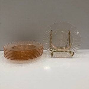 Puede incluir: Un conjunto de platos de vidrio rosa con borde ondulado y dise&ntilde;o en espiral en el centro. Un plato se exhibe en un soporte de metal dorado. Los platos est&aacute;n sobre una superficie blanca.