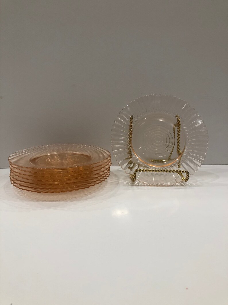Puede incluir: Un conjunto de platos de vidrio rosa con borde ondulado y dise&ntilde;o en espiral en el centro. Un plato se exhibe en un soporte de metal dorado. Los platos est&aacute;n sobre una superficie blanca.