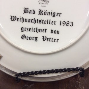 以下が含まれることがあります： 黒い文字で「Bad K&ouml;niger Weihnachtsteller 1983 gezeichnet von Georg Vetter」と書かれた白い磁器の皿。皿には、「Lang Ebrach Bavaria Germany」という文字と王冠のロゴがある。