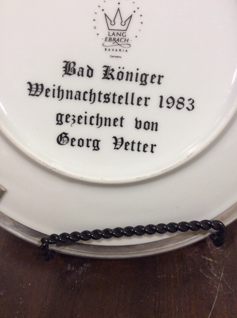 以下が含まれることがあります： 黒い文字で「Bad K&ouml;niger Weihnachtsteller 1983 gezeichnet von Georg Vetter」と書かれた白い磁器の皿。皿には、「Lang Ebrach Bavaria Germany」という文字と王冠のロゴがある。