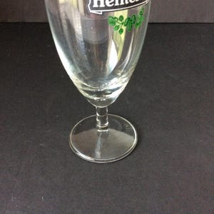 May include: A clear, stemmed beer glass with a wide bowl and a circular base. The glass has a logo with green accents at the top. The glass is empty and appears to be clean.