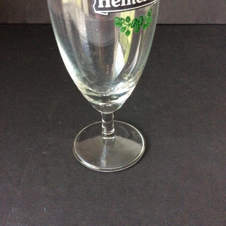 May include: A clear, stemmed beer glass with a wide bowl and a circular base. The glass has a logo with green accents at the top. The glass is empty and appears to be clean.