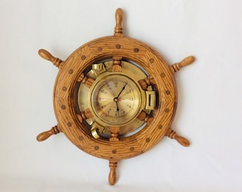 May include: A wooden ship's wheel clock with a brass clock face. The clock has a light brown wooden frame with six spokes and a brass clock face. The clock face has black numbers and hands. The clock is mounted on a white background.