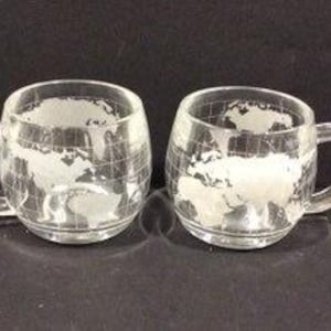 May include: Two clear glass mugs with handles, each etched with a world map design. The mugs have a rounded shape and sit on a dark surface. The world map design is in white, contrasting with the clear glass.