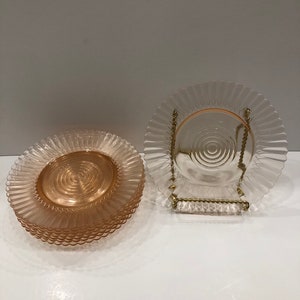 Puede incluir: Un conjunto de platos de vidrio rosa con borde ondulado y dise&ntilde;o espiral central. Un plato se exhibe en un soporte de metal dorado. Los platos tienen una est&eacute;tica vintage y son adecuados para servir alimentos.