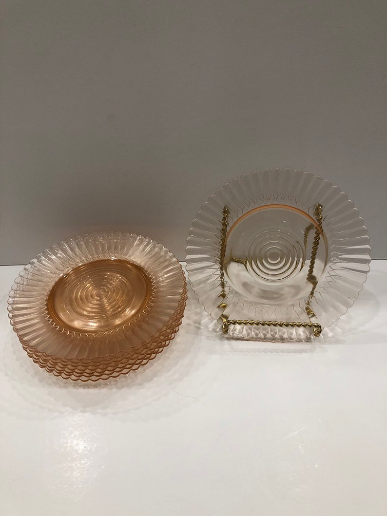 Puede incluir: Un conjunto de platos de vidrio rosa con borde ondulado y dise&ntilde;o espiral central. Un plato se exhibe en un soporte de metal dorado. Los platos tienen una est&eacute;tica vintage y son adecuados para servir alimentos.