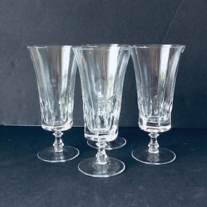 Può includere: Quattro flûte da champagne trasparenti dal design classico. I bicchieri hanno una coppa svasata, uno stelo dettagliato e una base rotonda. Le flûte sono realizzate in vetro trasparente e sono disposte su una superficie scura.