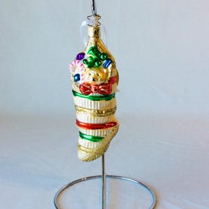 May include: A Christmas stocking ornament with a teddy bear, candy canes, and gifts. The ornament is cream-colored with gold glitter accents and red and green stripes. It hangs from a silver metal stand.