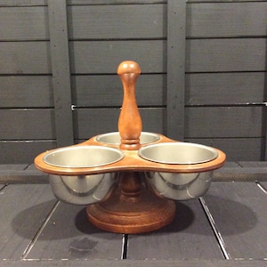 May include: A three-section serving dish featuring a central wooden handle and base with three removable stainless steel bowls. The wooden components have a medium brown finish, while the bowls are polished silver in color.