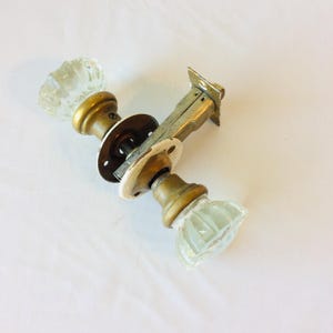May include: A vintage door knob set featuring two clear, fluted glass knobs attached to brass-toned metal bases. The assembly includes a central metal latch mechanism with a white circular mounting plate and a rectangular metal strike plate.