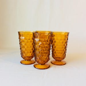 Op de afbeelding: Vier amberkleurige drinkglazen. Elk glas heeft een getextureerd, vierkant patroon en een voet. De glazen zijn in een groep gerangschikt en tonen hun vintage design. De glazen zijn gemaakt van glas.