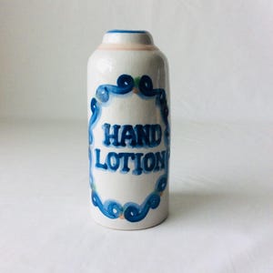 May include: A white ceramic hand lotion bottle with blue and green painted accents. The bottle has a blue and white decorative border around the words "HAND LOTION". The top of the bottle is light pink and blue.