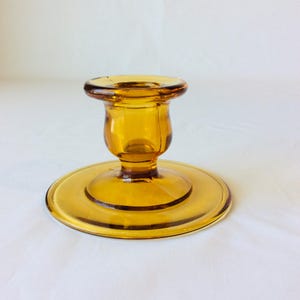 May include: Amber-colored glass candlestick holder with a round base and a flared top. The candlestick holder has a classic design and is made of transparent glass. The base is approximately 4 inches in diameter.