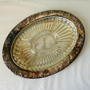 Può includere: Vassoio ovale con inserto in vetro trasparente e bordo in metallo color argento. Il bordo presenta un motivo decorativo a uva e foglie. L'inserto in vetro ha un design diviso con un'area circolare centrale e linee radiali.