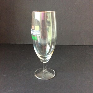 May include: A clear, stemmed beer glass with a gold rim. The glass features the Heineken logo in green and white. The glass is empty and sits on a dark surface.