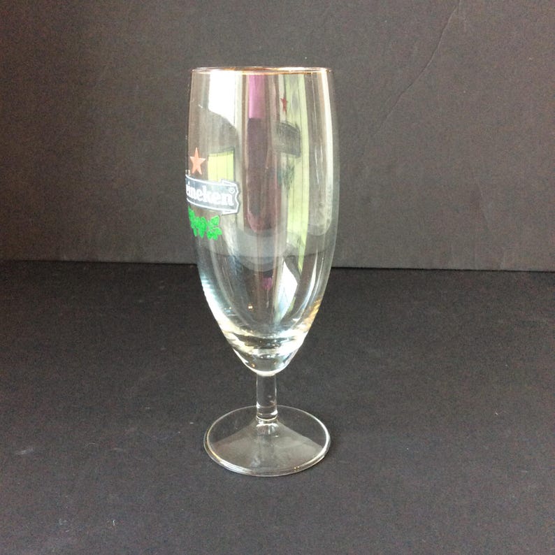 May include: A clear, stemmed beer glass with a gold rim. The glass features the Heineken logo in green and white. The glass is empty and sits on a dark surface.