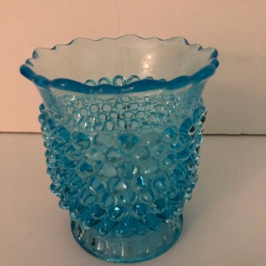 May include: A small, light blue glass vase with a textured, bubbled surface. The vase has a scalloped rim and a flared base.