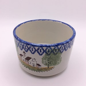 May include: A white ceramic bowl with a blue and white geometric pattern around the rim. The bowl has a painted scene of a small village with a church, trees, and houses.