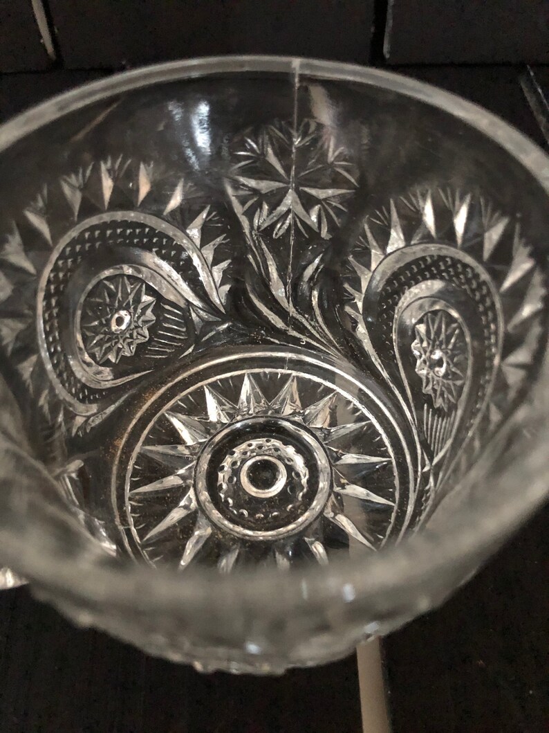 May include: Clear glass bowl with intricate starburst and swirl patterns. The bowl's design features detailed cuts and shapes, creating a textured surface. The bowl's rim is visible, and the overall aesthetic is vintage.