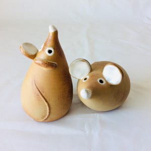 Puede incluir: Un par de figuritas de ratón de cerámica. Los ratones son marrones con detalles blancos y tienen un estilo caprichoso y de dibujos animados.