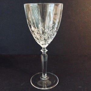 May include: A clear crystal wine glass with a decorative pattern on the bowl. The glass has a long stem and a round base. The glass is approximately 8 inches tall.
