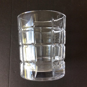 Op de afbeelding: Een doorzichtig drinkglas met een geometrisch rasterpatroon van verhoogde vierkante facetten over het buitenoppervlak. De beker heeft een cilindrische vorm met een gladde rand en een dikke, stevige bodem die het licht door de wanden breekt.