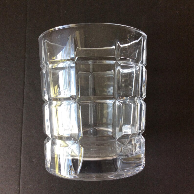 Op de afbeelding: Een doorzichtig drinkglas met een geometrisch rasterpatroon van verhoogde vierkante facetten over het buitenoppervlak. De beker heeft een cilindrische vorm met een gladde rand en een dikke, stevige bodem die het licht door de wanden breekt.