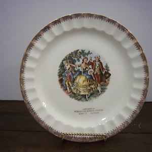 Pode incluir: Um prato decorativo branco com borda ondulada e borda dourada. O centro apresenta uma ilustração colorida de pessoas em trajes históricos. O texto na parte inferior diz "Compliments of Roelscher Service Station, Highway 75, Illinois."