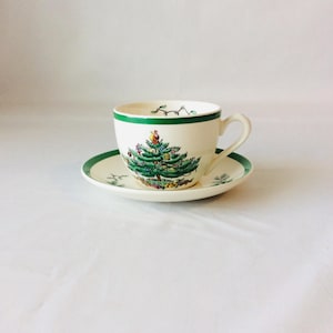 Può includere: Un set di tazza da tè e piattino in ceramica color crema con bordo verde e un'illustrazione centrale di un albero di Natale decorato con ornamenti colorati. La tazza ha un manico curvo e il piattino presenta dettagli verdi coordinati.