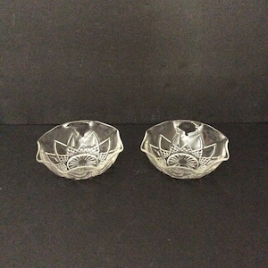May include: Two small, clear glass bowls featuring a pressed pattern with a starburst design at the center and a scalloped rim. The glass has a faceted texture that catches the light, creating a decorative appearance for these two matching dishes.