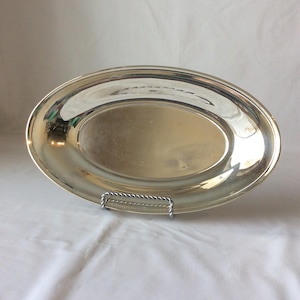 May include: An oval-shaped silver-toned serving platter with a smooth, reflective metallic surface and a raised rim. The platter is displayed resting on a small, silver-coloured wire stand that features a twisted rope-like texture design.