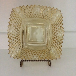 Puede incluir: Un plato cuadrado de vidrio color ámbar con un patrón de diamantes y un centro cuadrado. El plato se exhibe sobre un soporte de metal decorativo. Los bordes del plato tienen un diseño de dientes de sierra.