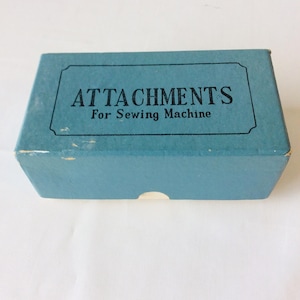 May include: A rectangular blue cardboard box with a black border and the text ATTACHMENTS For Sewing Machine printed in black on the top lid. The box features a small semi-circular cutout on the front edge and shows minor wear on the corners.