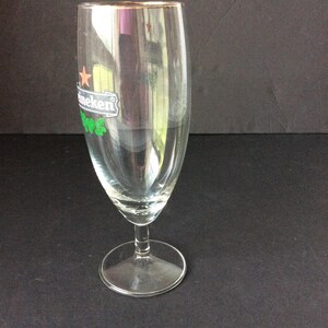 May include: A clear, stemmed beer glass with a gold rim. The glass features the Heineken logo in green and white. The glass is empty and sits on a dark surface.