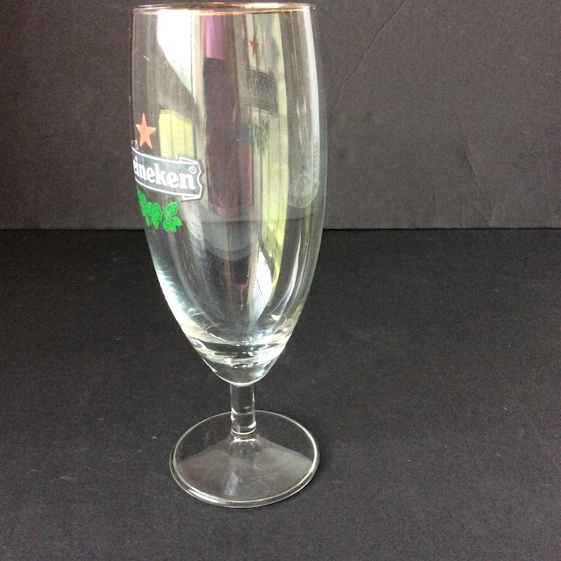 May include: A clear, stemmed beer glass with a gold rim. The glass features the Heineken logo in green and white. The glass is empty and sits on a dark surface.