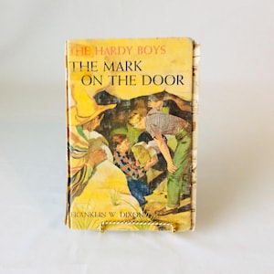 May include: Vintage hardcover book titled "The Hardy Boys: The Mark on the Door." The cover features an illustration of several figures in a landscape, with text in orange and black. The book is displayed on a gold-colored stand.