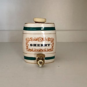 May include: A white ceramic sherry dispenser shaped like a barrel with a green and gold trim. The dispenser has a spout and says "SHERRY" on the front.