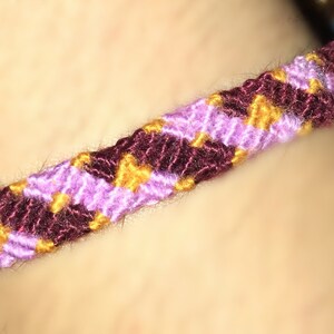 May include: A woven friendship bracelet with a chevron pattern. The bracelet is made with purple, burgundy, and gold threads. The design is visible, highlighting the texture and craftsmanship of the accessory.