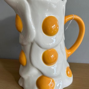 May include: A white ceramic jug with a quirky fried egg design. The jug has a bright orange handle and egg yolk details. This unique kitchenware item adds a playful touch to any table setting.