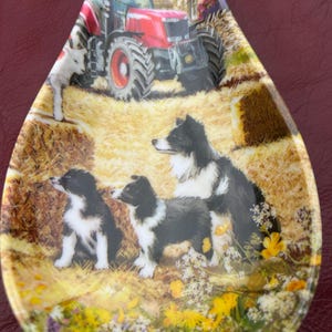 May include: A ceramic spoon rest with a rural design. It shows a red tractor, a white lamb, and several black and white Border Collies. The background includes a farmhouse and hay bales.