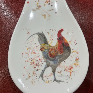 May include: White ceramic spoon rest featuring a rooster illustration. The rooster is red, yellow, and grey against a white background. The spoon rest has a speckled design with red, yellow, and brown paint splatters.