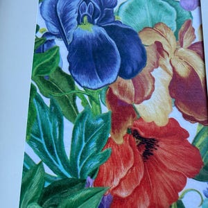 May include: A colourful botanical illustration featuring a prominent deep blue iris, a vibrant red poppy, and soft yellow-orange petals. The composition includes lush green leaves and stems, all set against a white background within a white frame.