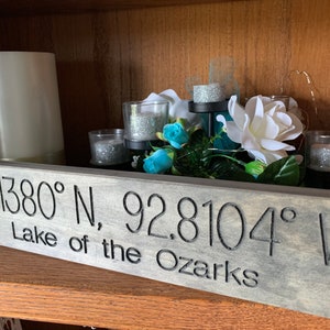May include: A wooden sign with the coordinates 38.1380° N, 92.8104° W and the text "Lake of the Ozarks" carved into it. The sign is sitting on a shelf with candles and flowers.