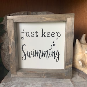 May include: A wooden framed sign with white background and black text that reads "just keep swimming" with black dots above the word swimming.