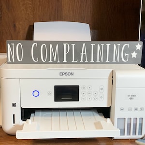May include: A white Epson ET-2760 printer with a sign above it that reads "NO COMPLAINING" in white letters on a gray background. The sign has two white stars on the right side.
