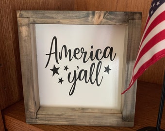 May include: A rustic wooden framed sign with the text "America y'all" in black lettering. There are three small stars on each side of the text. The sign is sitting on a wooden surface.