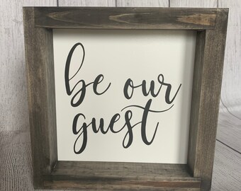 May include: A rustic wooden framed sign with the text "be our guest" in black cursive lettering on a white background.