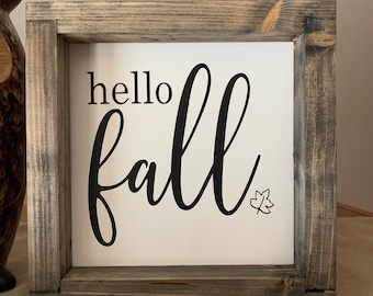 May include: A rustic wooden framed sign with the text "hello fall" in black script font. A small black leaf icon is in the bottom right corner of the sign.