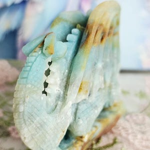 May include: A carved stone sculpture in shades of blue, tan, and white. The sculpture appears to be a stylized dragon or mythical creature. The details include carved scales and a textured surface. The sculpture is set against a soft, blurred background.